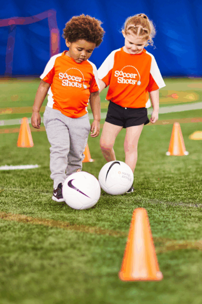 Two children in Soccer Shots jersey dribbling on turf